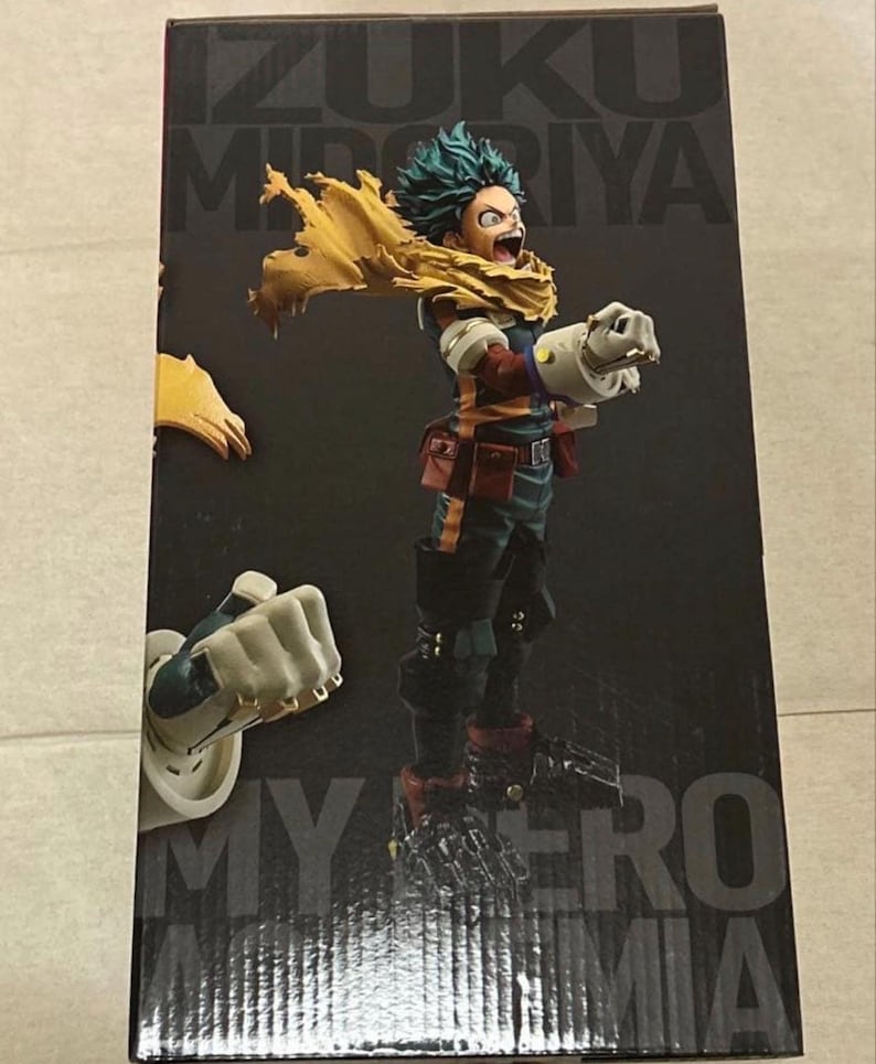May include: A collectible figure of Izuku Midoriya from My Hero Academia, depicted in a dynamic pose with a yellow scarf, dark green suit, and large white gauntlets. The figure is displayed on a black box featuring the character's name and series title.