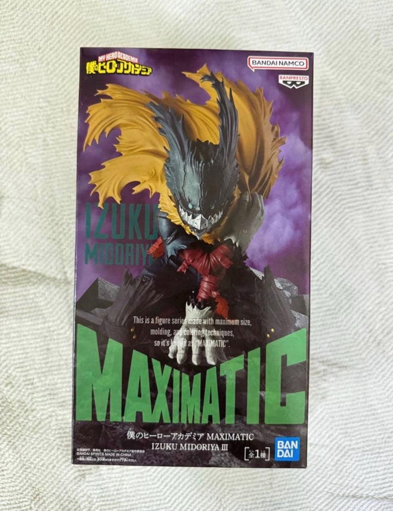 May include: A boxed MAXIMATIC figure of Izuku Midoriya from My Hero Academia. The figure has a detailed design with a black and yellow cape, and the box features green and purple text.