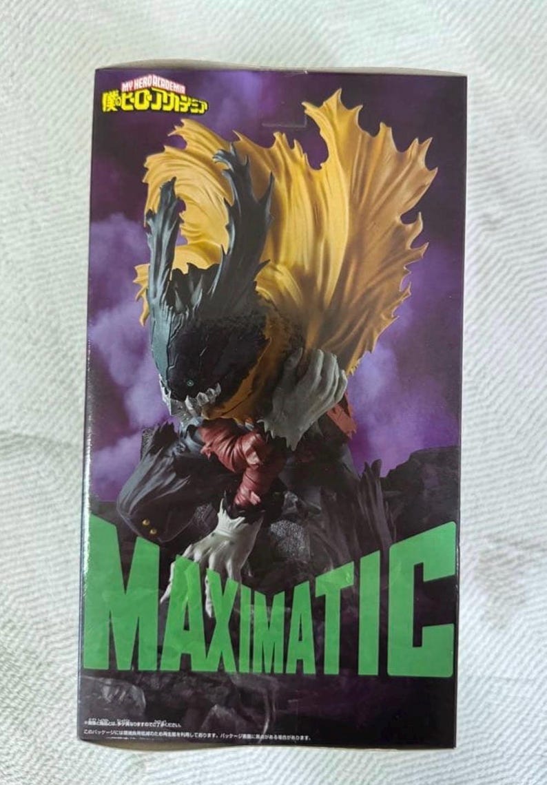 May include: A MAXIMATIC figure from the My Hero Academia series. The figure features a character with a black and yellow cape, gray hands, and a dark outfit. The box has the series logo and the word MAXIMATIC in green.