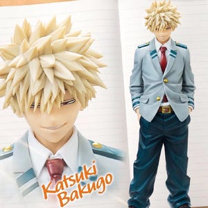 May include: A Katsuki Bakugo figure from the anime series. The figure has spiky blonde hair, a light blue blazer with green accents, a red tie, and dark blue pants. The figure is standing with hands in pockets. The name "Katsuki Bakugo" is printed on the notebook.