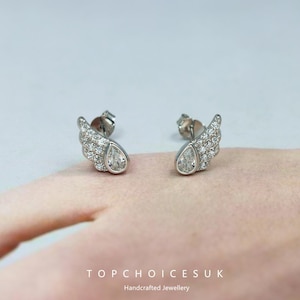 May include: Silver-tone stud earrings in a wing design. Each earring has a teardrop-shaped clear stone and is adorned with smaller, sparkling stones. The earrings are presented on a pink surface.