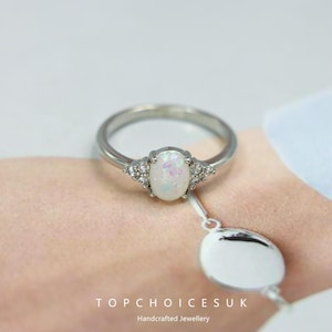 May include: A silver ring featuring an oval, iridescent opal center stone, flanked by small, clear accent stones. The ring band is thin and polished. The text "TOPCHOICESUK Handcrafted Jewellery" is visible at the bottom.