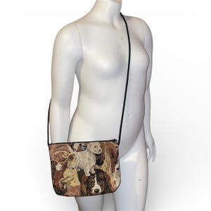 Peut inclure: Un sac bandoulière rectangulaire avec une sangle noire. Le sac présente un motif de tapisserie avec diverses races de chiens dans des tons de marron, beige et blanc. Le sac a une fermeture éclair noire.