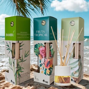 Può includere: Tre scatole di cartone rettangolari etichettate Bambu, Fruta Tropical e Pistachio, ciascuna con illustrazioni botaniche. Un diffusore a bastoncini in vetro con tappo bianco e bastoncini di legno è posizionato davanti con olio giallo.