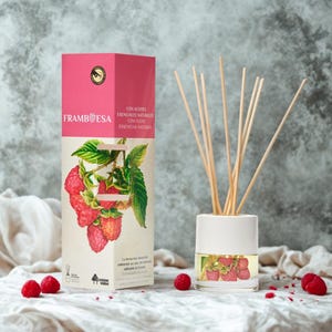 Puede incluir: Un difusor con aroma a frambuesa con un recipiente de cerámica blanca y varillas de madera. El empaque presenta la palabra "FRAMBESA" y una ilustración de frambuesa. Varias frambuesas frescas están dispersas.