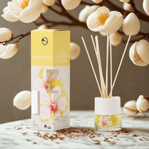 Może przedstawiać: Zestaw dyfuzora zapachowego Magnolia. Zestaw zawiera szklaną butelkę z kwiatowym wzorem, patyczki i żółte pudełko z nadrukiem "Magnolia". Pudełko ma również kwiatowy wzór.