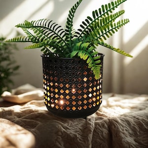 Op de afbeelding: Een zwarte metalen plantenpot met een ruitvormig patroon, gevuld met een levendige groene varen. De binnenkant van de pot gloeit warm, wat een gezellige sfeer creëert. De bloempot staat op een getextureerd, neutraal getint oppervlak.