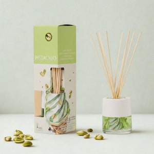 May include: A pistachio-scented reed diffuser set with a light green and white design. The set includes a glass bottle with reeds and a matching box with the word "PISTACHIO" on the front. Several pistachios are scattered around the set.