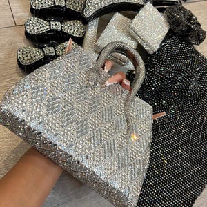 May include: A silver handbag covered in clear rhinestones with a geometric pattern, featuring a top handle. The set includes black hair clips with rhinestone bows, a small square rhinestone pouch, and a black rhinestone-covered fabric accessory.