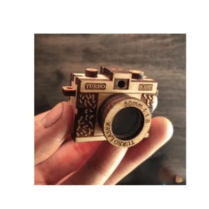 May include: A miniature wooden camera with the word "TURBO" on top. The lens is labeled "50mm 1:1.8" and "TURBO LASER". The camera has intricate cut-out designs on the sides and is held in a hand.