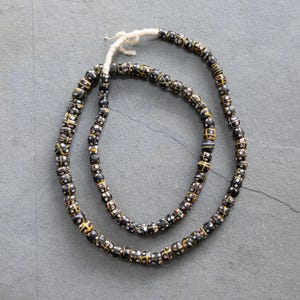 Puede incluir: Un largo collar de cuentas de vidrio negro ensartadas en un cordón blanco. Cada cuenta presenta un patrón decorativo de líneas amarillas entrecruzadas que forman una cuadrícula, con pequeños puntos blancos y rosas dentro de los cuadrados formados.
