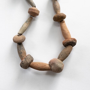 May include: A necklace featuring a series of alternating terracotta-colored clay beads in cylindrical and flattened disc shapes. The beads have a porous, earthy texture with variations in brown, tan, and grey tones, strung together on a cord.