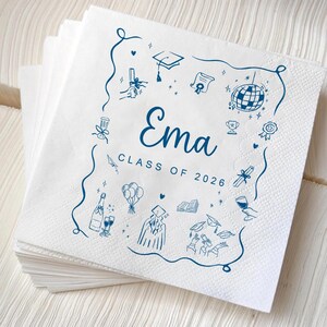 May include: A pile of white paper napkins featuring a blue illustrated border. The napkins are decorated with graduation-themed doodles and the name "Ema" with "Class of 2026" in a script font.