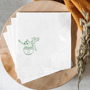 May include: White paper napkins with a green illustration of a woman in a pot of coins, with the text "Vale's 26th Birthday". The napkins are stacked on a wooden tray, with a yellow cloth and dried plants in the background.