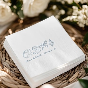 May include: A stack of white paper napkins with a blue seashell design and the text "Olivia's Bachelorette / Charleston, Sc". The napkins are on a woven basket.