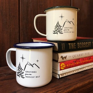 Puede incluir: Dos tazas de esmalte blanco con bordes azules y verdes. Ambas tazas presentan una ilustración en blanco y negro de un pino y montañas con el texto "Greetings from the Borscht Belt". Las tazas están apiladas sobre una pila de libros.