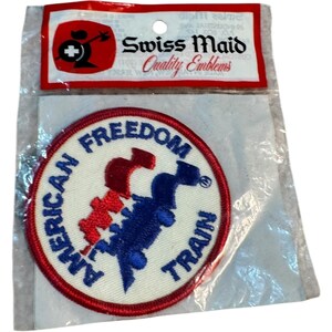 Pode incluir: Um patch circular com as palavras "AMERICAN FREEDOM TRAIN" em texto azul. O patch apresenta um design de comboio vermelho e azul sobre um fundo branco, com uma borda vermelha. O patch é embalado com uma etiqueta "Swiss Maid Quality Emblems".