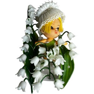 May include: A vintage figurine of a small doll with yellow hair and a white bonnet, surrounded by white lily of the valley flowers and green leaves. The doll is wearing a green outfit. The figurine is set against a white background.