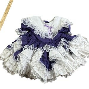 May include: A vintage purple and white dress with a white lace collar and ruffled skirt. The dress features a purple bow and lace trim, creating a classic, elegant look. The dress is likely for a child.