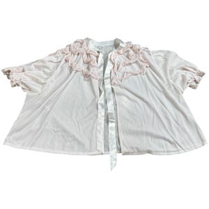 Könnte beinhalten: Ein vintage, hellrosa Bademantel mit Rüschenkragen und Manschetten. Der Bademantel hat einen Satin-Bindverschluss und zarte rosa Spitzentrimm. Die Ärmel sind kurz und der Stoff scheint ein leichtes, fließendes Material zu sein.
