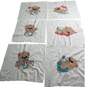 May include: A set of six white fabric squares featuring cross-stitched teddy bear designs. Each square displays a brown bear in different activities, including playing with a ball, holding a rattle, sitting with blocks, and drinking from a bottle.