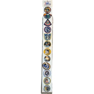 May include: A strip of Apollo mission patches, featuring various designs and colors. The patches are round and display mission emblems, text, and imagery. The top of the strip has the text "APOLLO EMBLEMS" and "KENNEDY SPACE CENTER".