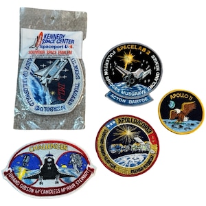 May include: A collection of embroidered space patches, including a Kennedy Space Center souvenir, a Spacelab 2 patch, an Apollo 11 patch with an eagle, a Challenger patch, and an Apollo/Soyuz patch. The patches feature colorful designs and text.