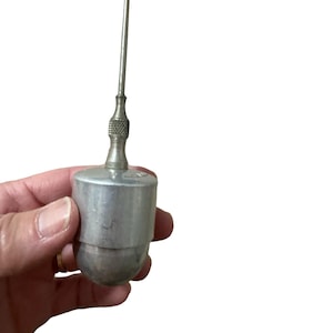 May include: A vintage metal tool with a cylindrical body and a long, thin, pointed metal rod extending from the top. The tool has a textured grip and a rounded base. The tool is held in a hand against a white background.