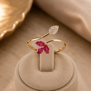 May include: A gold bypass ring featuring a leaf-shaped accent set with small clear stones and a cluster of three marquise-cut deep pink stones. The band is polished gold, creating a delicate open design that curves around the finger.