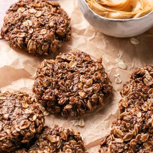 Op de afbeelding: Verschillende ronde, donkerbruine no-bake koekjes gemaakt van havermout en cacao, met een textuurrijk en klonterig oppervlak. Een klein wit keramieken kommetje gevuld met romige lichtbruine pindakaas staat bij de koekjes.