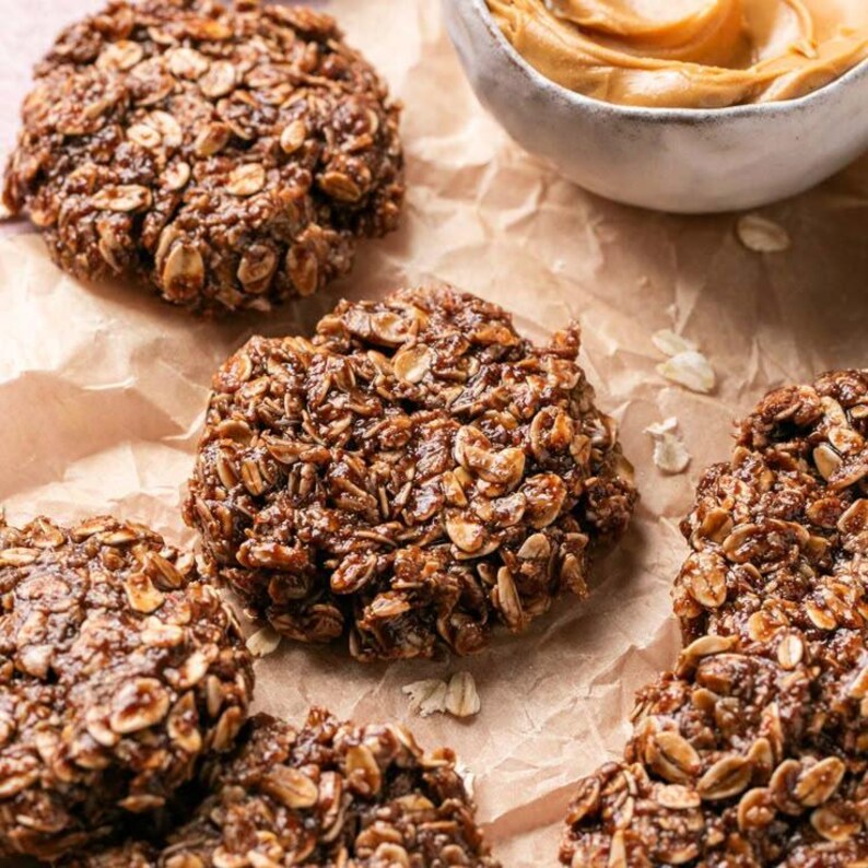 Op de afbeelding: Verschillende ronde, donkerbruine no-bake koekjes gemaakt van havermout en cacao, met een textuurrijk en klonterig oppervlak. Een klein wit keramieken kommetje gevuld met romige lichtbruine pindakaas staat bij de koekjes.