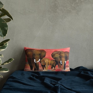 May include: Rectangular decorative pillow featuring a painting of a family of elephants. The elephants are brown with white tusks, set against a red and orange background. The pillow is placed on a dark blue blanket.