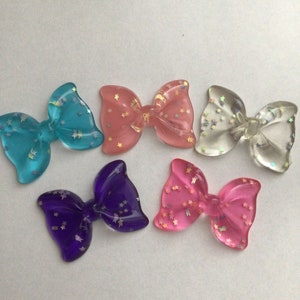 May include: Five resin bows with glitter embedded inside. The bows are in the colors blue, pink, clear, purple, and pink.