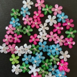 May include: Assortment of colorful plastic flower embellishments with rhinestones in the center. The flowers are in shades of pink, blue, green, and white.