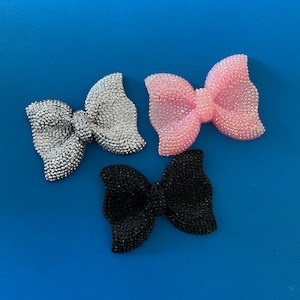 May include: Three sparkly hair bows in silver, pink, and black. The bows are arranged in a triangle shape on a blue background.