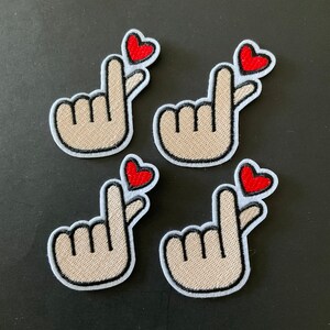 May include: Four white embroidered patches with a hand gesture and a red heart. The hand gesture is a Korean heart sign, also known as the mini heart or finger heart.