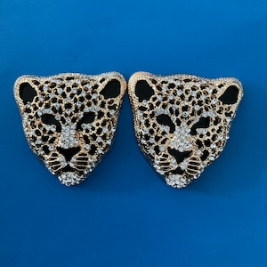 May include: A pair of gold-tone earrings with a black and gold leopard design. The earrings are adorned with clear rhinestones.
