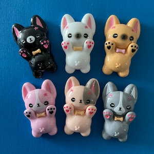 May include: Six small, colorful, cartoon dog figurines. Each dog is holding a bone in its paws and has a pink bow tie. The dogs are in various colors, including black, white, tan, pink, and gray.