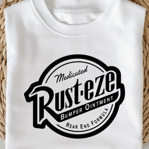 May include: White sweatshirt featuring a black and white graphic. The graphic is a circular design with the words "Medicated Rust-eze Bumper Ointment Rear End Formula" in a vintage style.