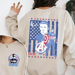 May include: Beige sweatshirt featuring a Captain America graphic. The design includes an American flag, the character's image, shield, and the text "1941". A smaller graphic of Captain America is on the lower left.