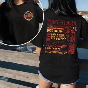 May include: Black t-shirt featuring the text "TEAM STARK" on the front and a graphic of Tony Stark with various quotes and phrases on the back. The design includes the words "TONY STARK", "I AM IRON MAN", and "STARK INDUSTRIES".