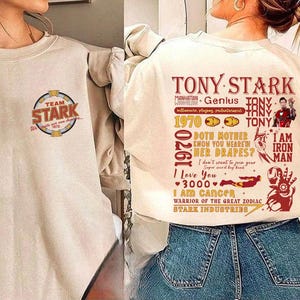 May include: Cream-colored sweatshirt with "TEAM STARK" logo and text. The back features a graphic with "TONY STARK" and other text, including "I AM IRON MAN" and "STARK INDUSTRIES".