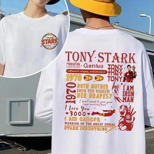 May include: White t-shirt featuring a circular "TEAM STARK" logo on the front. The back displays a graphic with the text "TONY STARK", "I AM IRON MAN", and other related phrases and illustrations in red and gold.