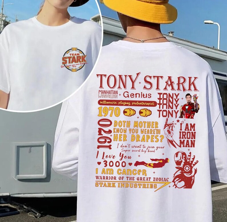 May include: White t-shirt featuring a circular "TEAM STARK" logo on the front. The back displays a graphic with the text "TONY STARK", "I AM IRON MAN", and other related phrases and illustrations in red and gold.