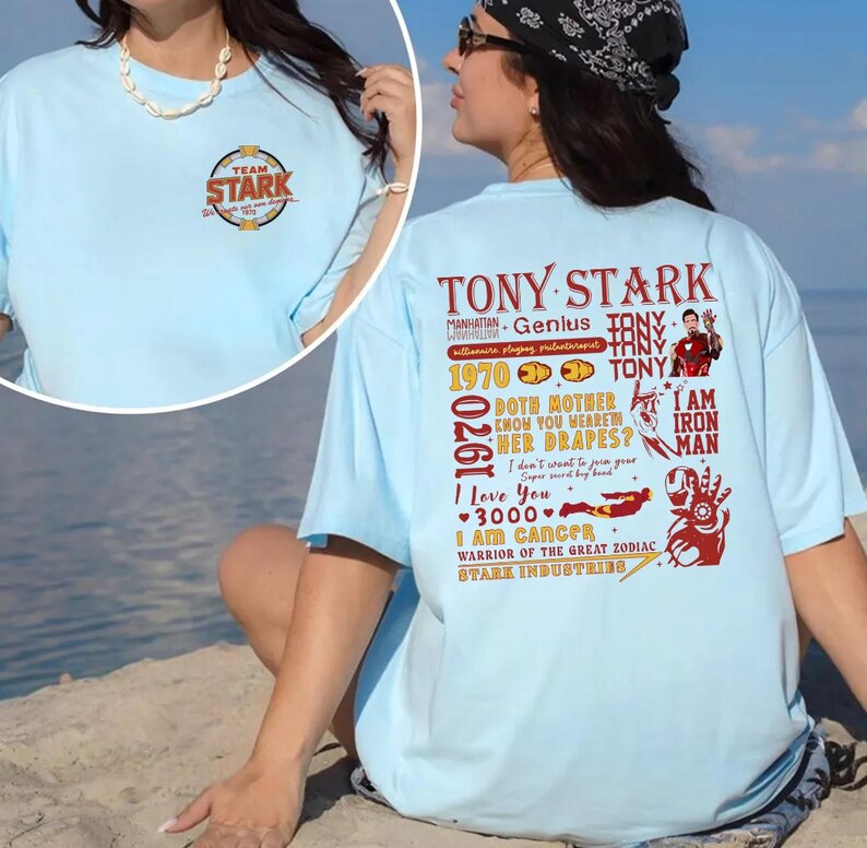 May include: Light blue t-shirt with "TEAM STARK" logo on the front. The back features a graphic with the text "TONY STARK" and other text and graphics in red. The shirt is worn by a person.