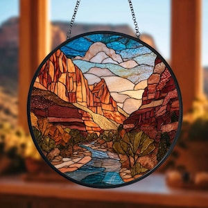 May include: Round stained glass window depicting a canyon landscape with a river. The artwork features shades of orange, red, brown, blue, and green. The glass is framed in black and hangs from a chain.