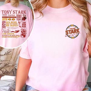 May include: A light pink t-shirt featuring the text "TEAM STARK" in a circular design on the chest. The back of the shirt has a graphic with the words "TONY STARK" and other text and images.