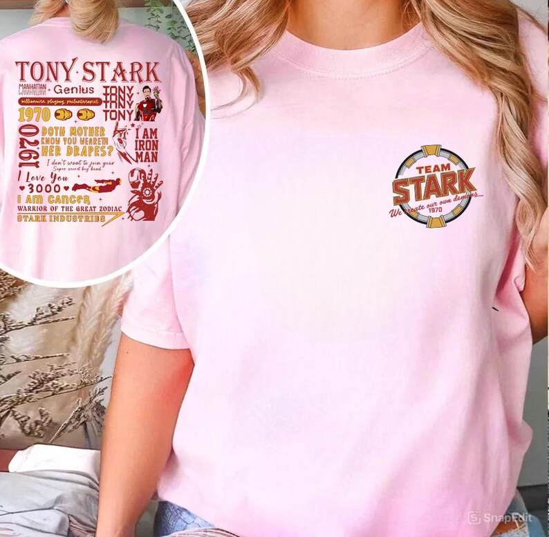 May include: A light pink t-shirt featuring the text "TEAM STARK" in a circular design on the chest. The back of the shirt has a graphic with the words "TONY STARK" and other text and images.