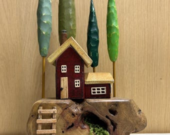 May include: A decorative wooden sculpture featuring a small red house with a tan roof, positioned on a natural wood base. Four carved trees in shades of green stand behind the house. A small wooden ladder leans against the base, which includes a hollow opening containing green moss.