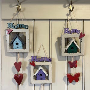 May include: Three decorative wooden birdhouse ornaments hanging from hooks. Each ornament features a small birdhouse inside a white frame with the word "Home" above. Red hearts and butterflies dangle below. The birdhouses are painted blue, green, and purple.